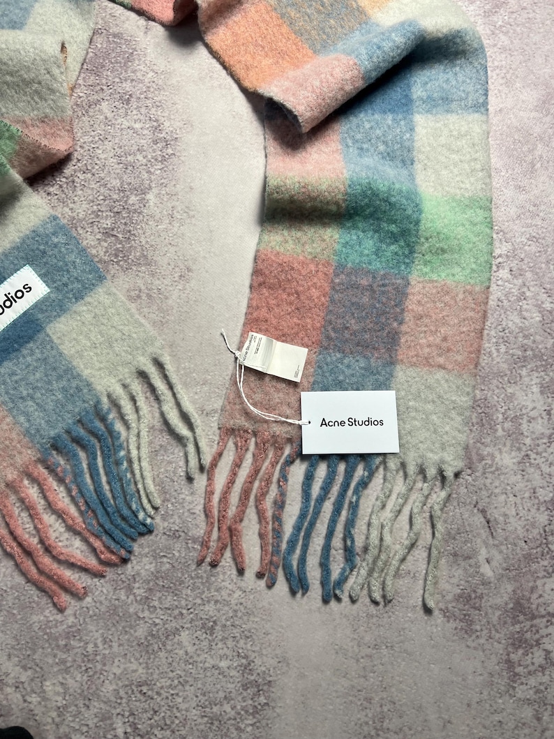 May include: A colorful, checkered scarf in pastel shades of pink, blue, green, and peach. The scarf has a fringed edge and a tag that reads "Acne Studios". The scarf is made of a soft, woven material.