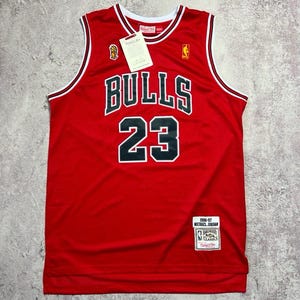 Puede incluir: Una camiseta de baloncesto roja de los Chicago Bulls con el número 23 en negro y borde blanco. El frente muestra la palabra BULLS en letras negras. Incluye una etiqueta de Mitchell & Ness, un logotipo de la NBA y un parche de Michael Jordan 1996-97.