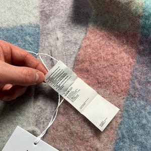 May include: A multi-colored scarf with a tag attached. The scarf features a patchwork design in shades of pink, blue, green, and gray. The tag is white with black text and the brand name "Acne Studios".