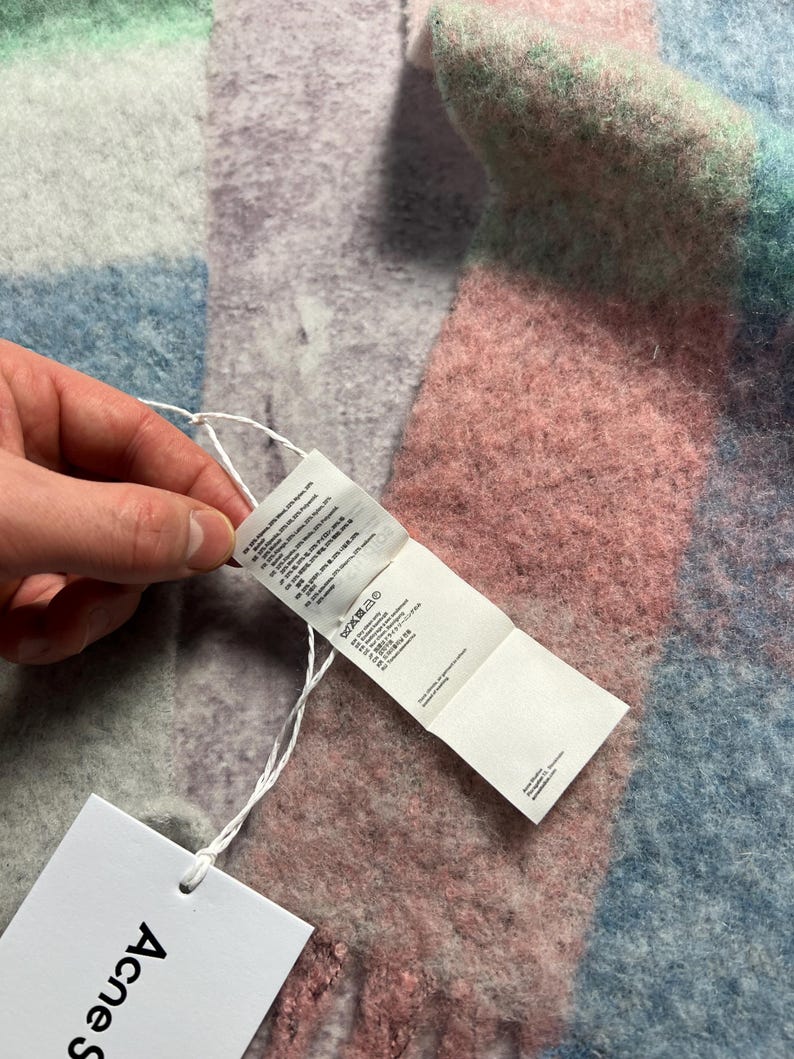 May include: A multi-colored scarf with a tag attached. The scarf features a patchwork design in shades of pink, blue, green, and gray. The tag is white with black text and the brand name "Acne Studios".