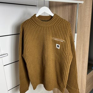 May include: A brown ribbed knit sweater with a mock neck and long sleeves. The sweater features a small pocket on the chest with a zipper and a black and white logo patch. The sweater is hanging on a white hanger.