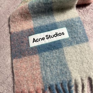 May include: A colorful scarf with a plaid pattern in shades of pink, blue, green, and gray. The scarf has a fringed edge and a white label that reads "Acne Studios".