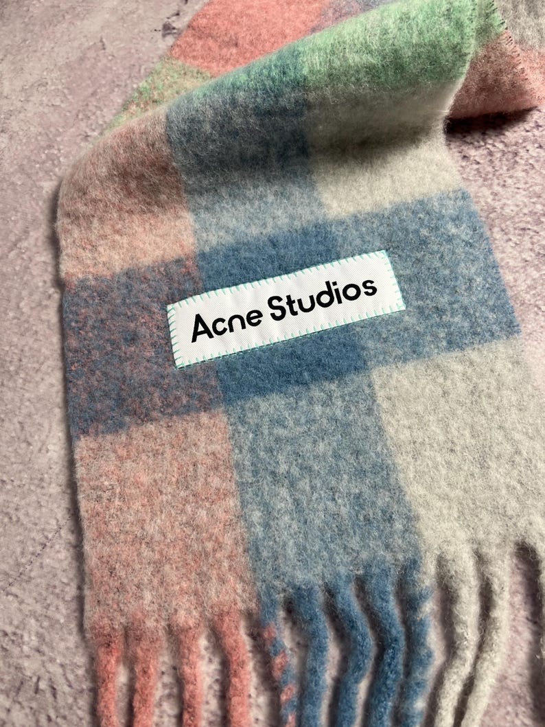 May include: A colorful scarf with a plaid pattern in shades of pink, blue, green, and gray. The scarf has a fringed edge and a white label that reads "Acne Studios".