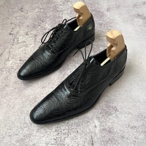 May include: Pair of black leather dress shoes with a textured pattern and lace-up closure. Wooden shoe trees are inserted inside each shoe. The shoes are displayed on a neutral background.