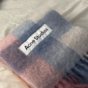 May include: A soft, fringed scarf in a color block design featuring shades of pink, blue, and gray. The scarf has the text "Acne Studios" on a white label.