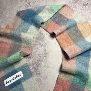 May include: A colorful, checkered scarf in pastel shades of pink, blue, green, and white. The scarf has a fringed edge and a tag that reads "Acne Studios". The scarf is made of a soft, fuzzy material.