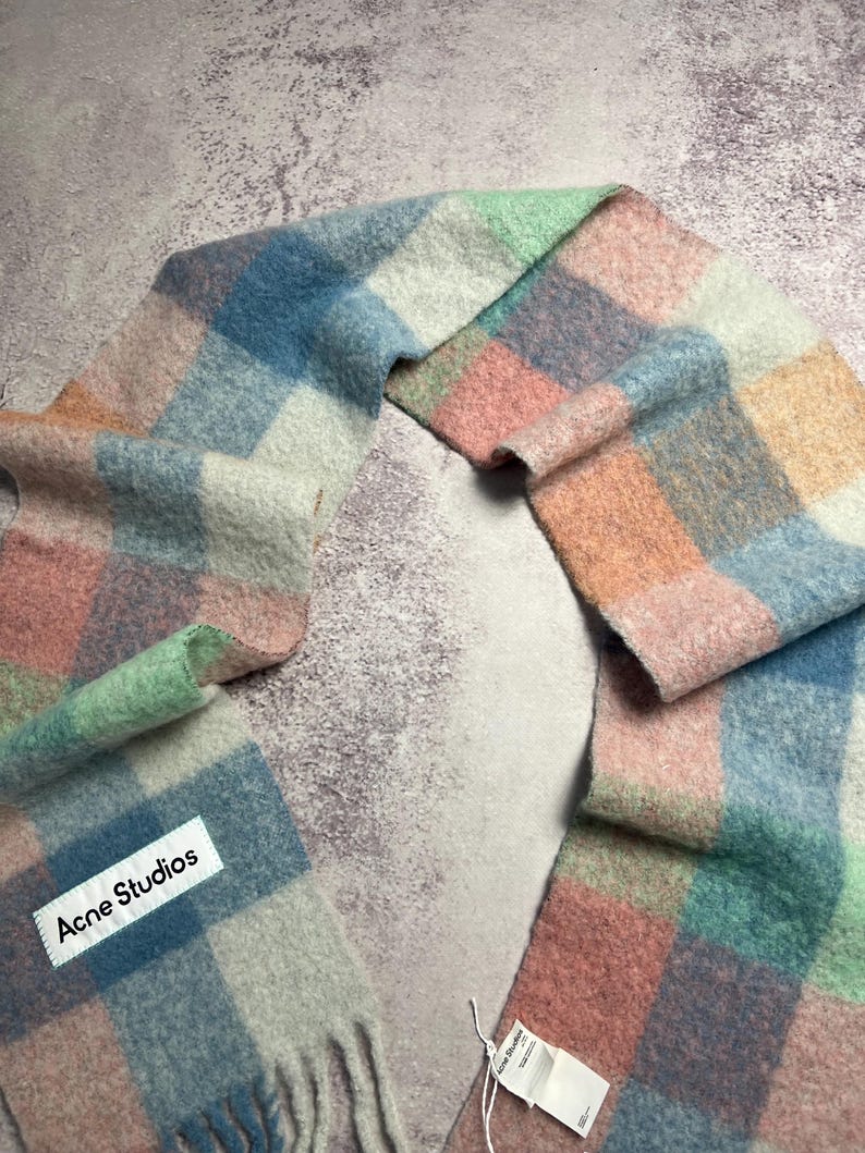 May include: A colorful, checkered scarf in pastel shades of pink, blue, green, and white. The scarf has a fringed edge and a tag that reads "Acne Studios". The scarf is made of a soft, fuzzy material.