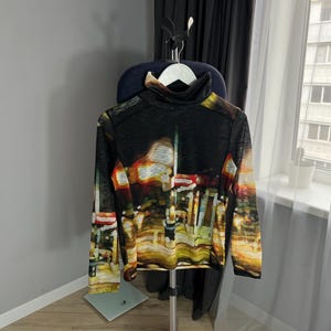 May include: A black turtleneck sweater with a colorful abstract print of a city skyline. The sweater is hanging on a hanger.