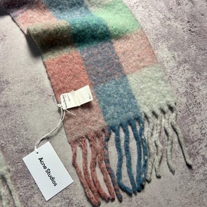 May include: A colorful scarf with a checkered pattern in shades of pink, blue, green, and white. The scarf has a fringed edge and a tag that reads "Acne Studios". The scarf is made of a soft, fuzzy material.