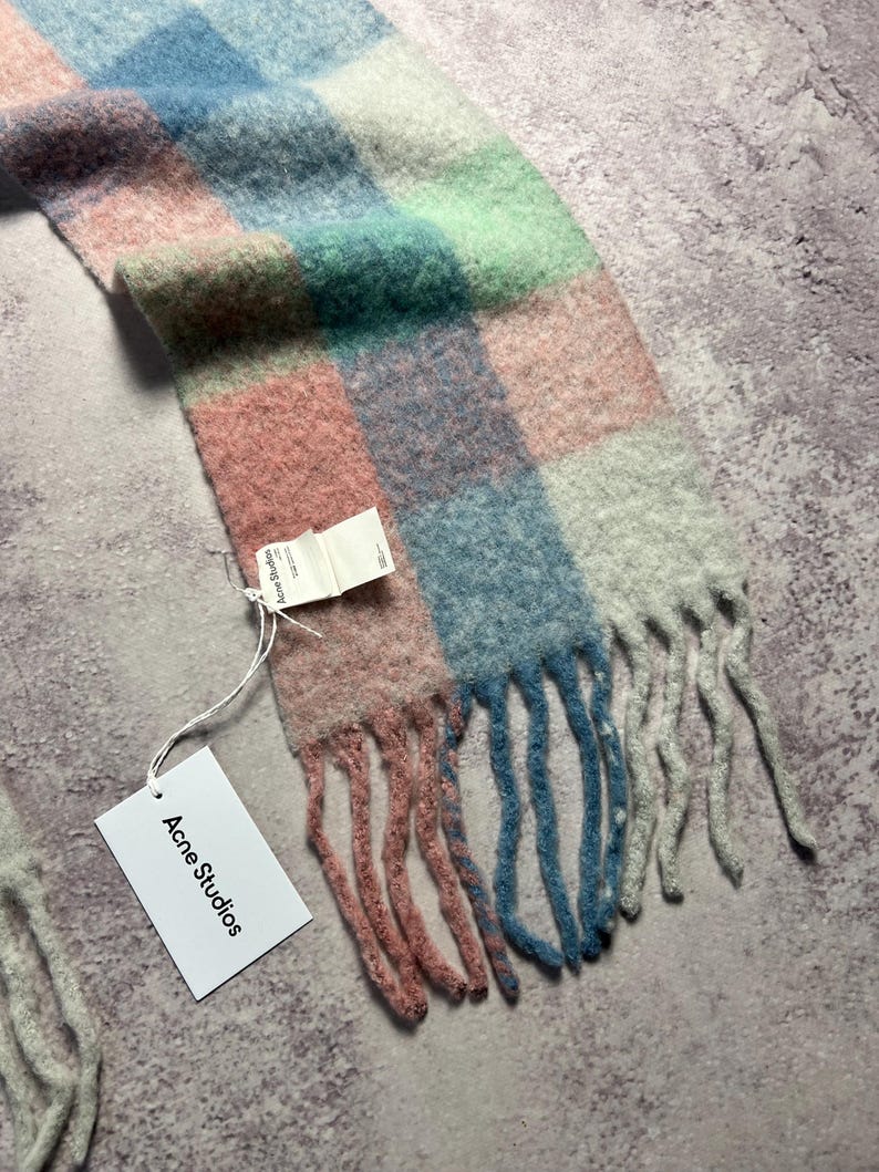 May include: A colorful scarf with a checkered pattern in shades of pink, blue, green, and white. The scarf has a fringed edge and a tag that reads "Acne Studios". The scarf is made of a soft, fuzzy material.