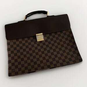 May include: A brown and dark brown checkered briefcase featuring a solid dark brown leather flap, a top handle with gold-tone hardware, and a central gold-tone metal clasp engraved with the Louis Vuitton brand name on the front surface.