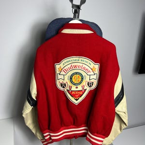 May include: Red and cream-colored varsity jacket with the Budweiser logo on the back. The jacket features a red body, cream-colored sleeves, and a white and red striped hem. The Budweiser logo includes the text "King of Bee".