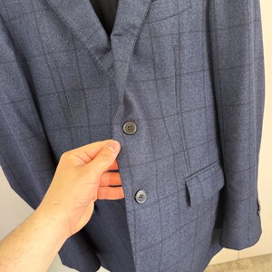 May include: A navy blue blazer featuring a dark charcoal windowpane check pattern. The garment includes a notched lapel, two dark buttons on the front closure, a chest pocket, and two flap pockets on the lower front section of the jacket.