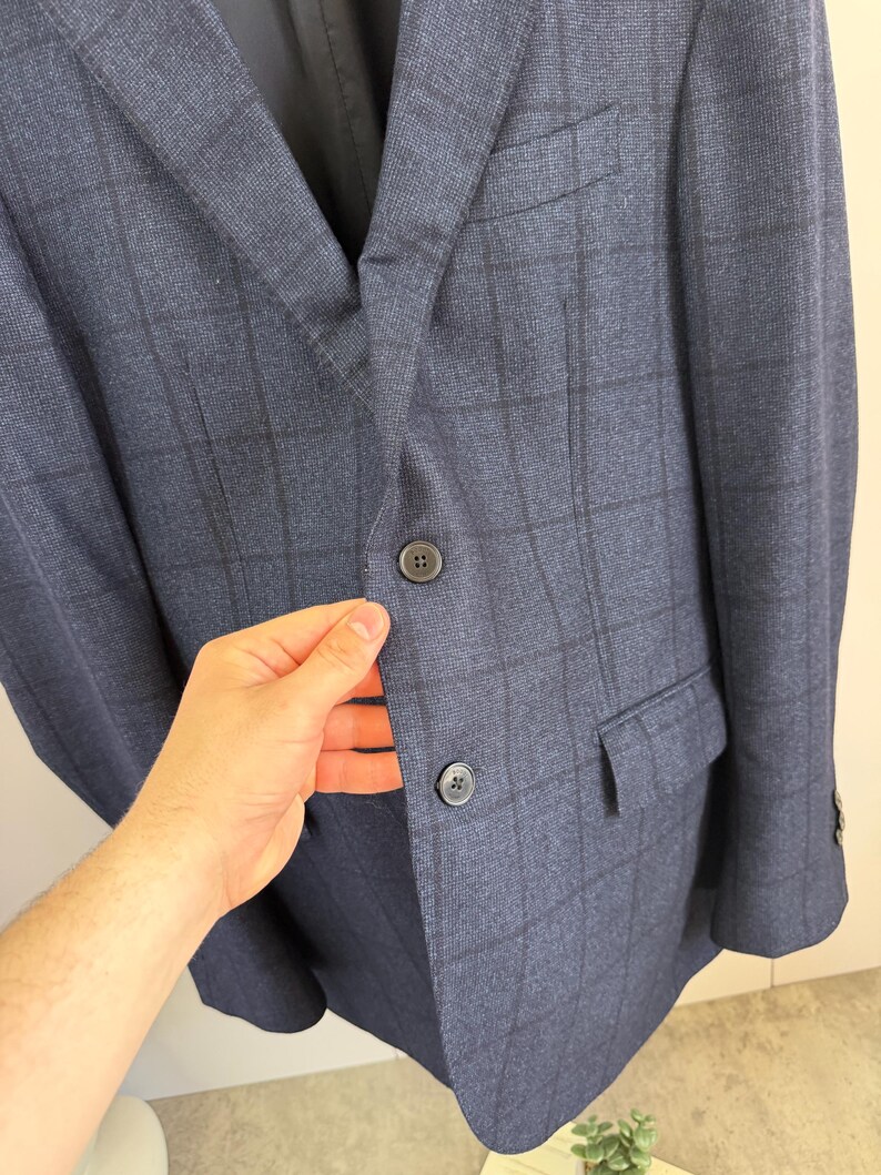 May include: A navy blue blazer featuring a dark charcoal windowpane check pattern. The garment includes a notched lapel, two dark buttons on the front closure, a chest pocket, and two flap pockets on the lower front section of the jacket.