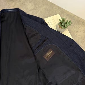 May include: A dark blue blazer with a subtle windowpane check pattern. The interior is fully lined in black fabric and features a rectangular label with the text Vitale Barberis Canonico 1663. A small Made in Italy tag is visible at the collar.