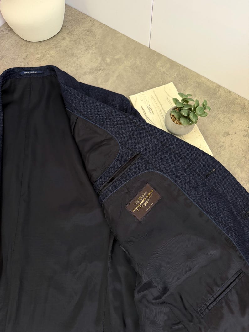 May include: A dark blue blazer with a subtle windowpane check pattern. The interior is fully lined in black fabric and features a rectangular label with the text Vitale Barberis Canonico 1663. A small Made in Italy tag is visible at the collar.