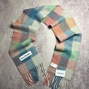 May include: A colorful, checkered scarf in pastel shades of blue, pink, green, and orange. The scarf has a fringed edge and a tag that reads "Acne Studios". The scarf is made of a soft, woven material.