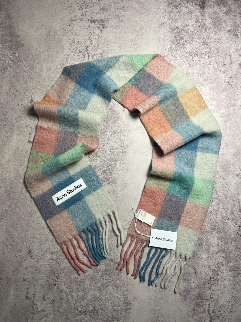 May include: A colorful, checkered scarf in pastel shades of blue, pink, green, and orange. The scarf has a fringed edge and a tag that reads "Acne Studios". The scarf is made of a soft, woven material.
