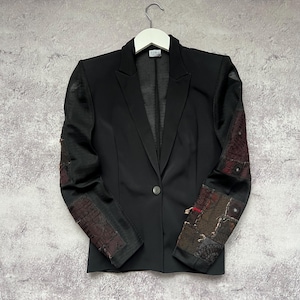 May include: Black blazer with a single-button closure and decorative patchwork on the sleeves. The blazer is hanging on a white hanger against a gray textured background. The sleeves feature a mix of dark red, brown, and black patches.
