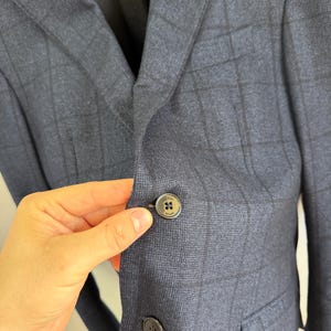 May include: A navy blue blazer featuring a dark charcoal windowpane check pattern. The garment has a notched lapel, a chest pocket, and two front flap pockets. A dark button with the text Boggi Milano is visible on the front closure.
