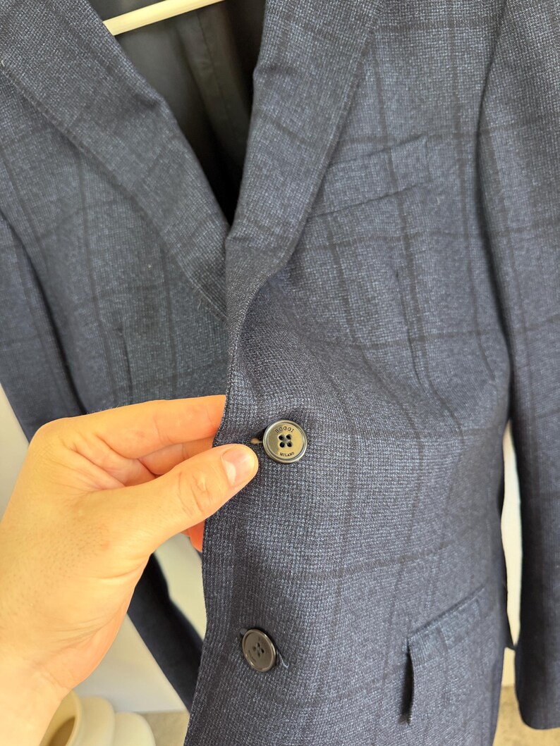 May include: A navy blue blazer featuring a dark charcoal windowpane check pattern. The garment has a notched lapel, a chest pocket, and two front flap pockets. A dark button with the text Boggi Milano is visible on the front closure.