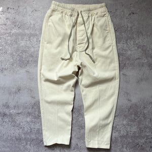 May include: Cream-colored casual pants with an elastic waistband and drawstring. The pants have a relaxed fit and are made of a soft, lightweight fabric. The pants have two front pockets and a single back pocket. The pants are suitable for everyday wear.