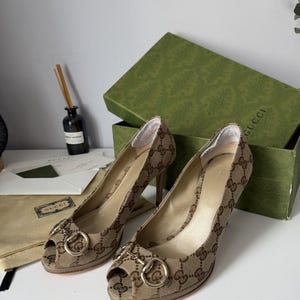 Peut inclure: Une paire d'escarpins peep-toe en toile beige et marron avec le motif monogramme Gucci et un détail mors en métal doré sur le devant. Les chaussures ont un talon haut et sont présentées avec une boîte verte de la marque et un pochon beige.