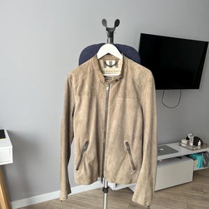 May include: A tan suede jacket with a zip-up front and two zippered pockets. The jacket is hanging on a white hanger attached to a clothing rack. The jacket has a collar and a brand label.