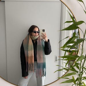 May include: A multi-colored scarf with a fringed edge. The scarf features a plaid pattern in shades of green, pink, and tan. The person in the image is wearing sunglasses and a black jacket. The image is reflected in a round mirror.