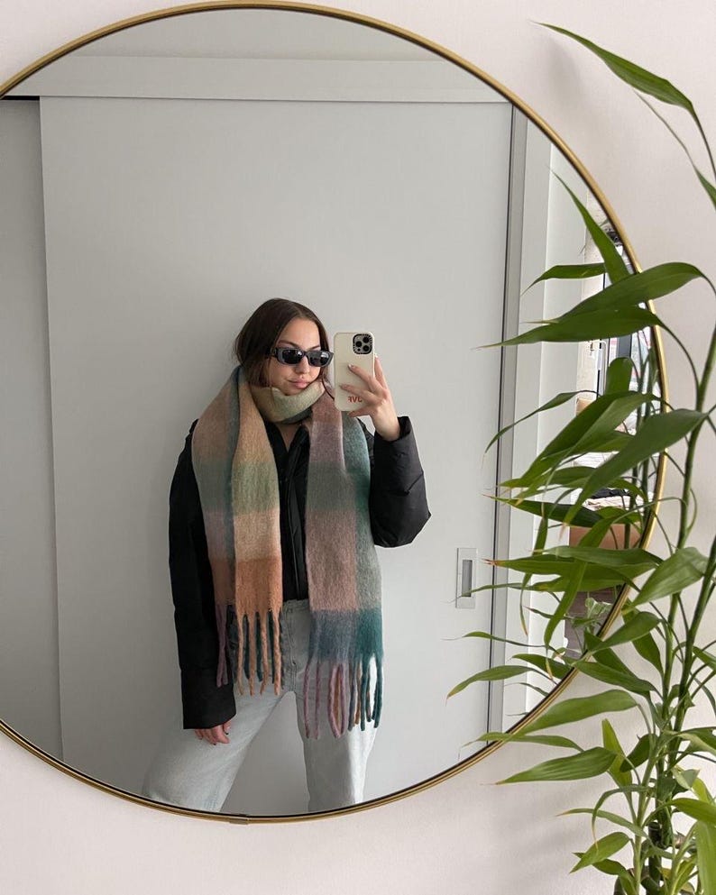 May include: A multi-colored scarf with a fringed edge. The scarf features a plaid pattern in shades of green, pink, and tan. The person in the image is wearing sunglasses and a black jacket. The image is reflected in a round mirror.