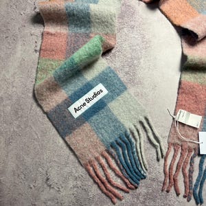 May include: A colorful scarf with a plaid pattern in shades of pink, blue, green, and gray. The scarf has a fringed edge and a white label that reads "Acne Studios".