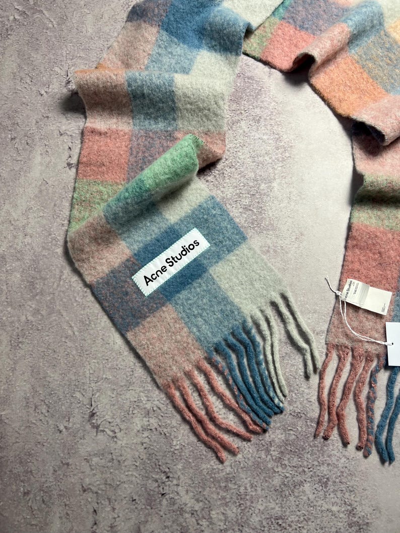 May include: A colorful scarf with a plaid pattern in shades of pink, blue, green, and gray. The scarf has a fringed edge and a white label that reads "Acne Studios".