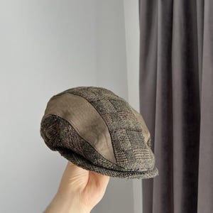 May include: A flat cap featuring a combination of brown and grey tweed fabric with a textured plaid pattern and solid brown panels on the sides. The cap has a rounded crown and a short, stiff brim extending from the front of the headwear.