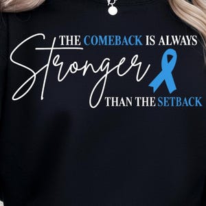 May include: Black sweatshirt with the text "The Comeback Is Always Stronger Than The Setback" in white script. A blue ribbon graphic is included. The sweatshirt is a casual, comfortable style.