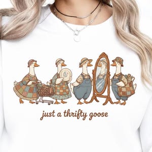 Pode incluir: Camisola branca com uma ilustração fantasiosa de gansos vestidos com roupas vintage. Os gansos são retratados com chapéus, aventais e macacões, alguns segurando itens como uma cesta e um espelho. O texto "just a thrifty goose" está impresso abaixo.
