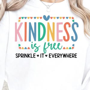 以下が含まれることがあります： カラフルな文字で「KINDNESS is free」と書かれた白いスウェットシャツ。「SPRINKLE IT EVERYWHERE」というフレーズが下に書かれています。デザインには、ハートやその他の装飾要素が様々な色で含まれています。