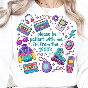May include: White sweatshirt with a colorful 1990s-themed graphic. The design includes a boombox, cassette tapes, roller skates, a handheld game console, and the text "please be patient with me, I'm from the 1900's."