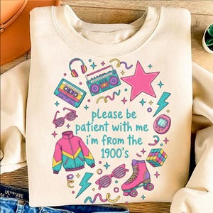 May include: Cream-colored sweatshirt with a colorful 1990s-themed graphic. The design includes a boombox, cassette tape, sunglasses, a jacket, roller skates, and the text "please be patient with me I'm from the 1900's."