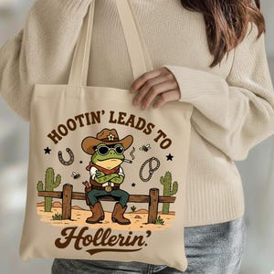 May include: Beige tote bag featuring a cartoon frog dressed as a cowboy, wearing a hat and sunglasses, smoking a cigarette, and sitting on a wooden fence. The text "Hootin' Leads to Hollerin'" is printed on the bag.