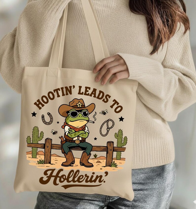 May include: Beige tote bag featuring a cartoon frog dressed as a cowboy, wearing a hat and sunglasses, smoking a cigarette, and sitting on a wooden fence. The text "Hootin' Leads to Hollerin'" is printed on the bag.
