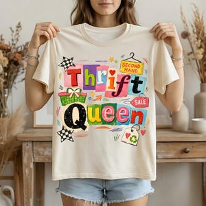 May include: Cream-colored t-shirt with the words "Thrift Queen" in colorful block letters. The design includes a shopping basket, stars, and a tag that says "Second Hand Sale".
