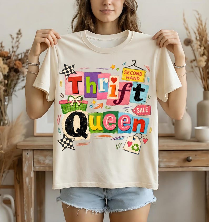 May include: Cream-colored t-shirt with the words "Thrift Queen" in colorful block letters. The design includes a shopping basket, stars, and a tag that says "Second Hand Sale".