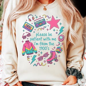 May include: Beige sweatshirt with a colorful 1900s-themed graphic. The design includes a pink star, roller skates, a boombox, and the text "please be patient with me I'm from the 1900's". Other elements include a cassette tape, sunglasses, and a jacket.