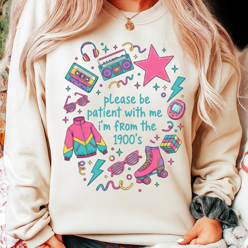 May include: Beige sweatshirt with a colorful 1900s-themed graphic. The design includes a pink star, roller skates, a boombox, and the text "please be patient with me I'm from the 1900's". Other elements include a cassette tape, sunglasses, and a jacket.