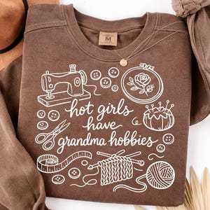 May include: A brown sweatshirt with white graphic illustrations of sewing tools, buttons, and knitting supplies. The text "hot girls have grandma hobbies" is written in a cursive font. A gold necklace and a brown hat are also visible.