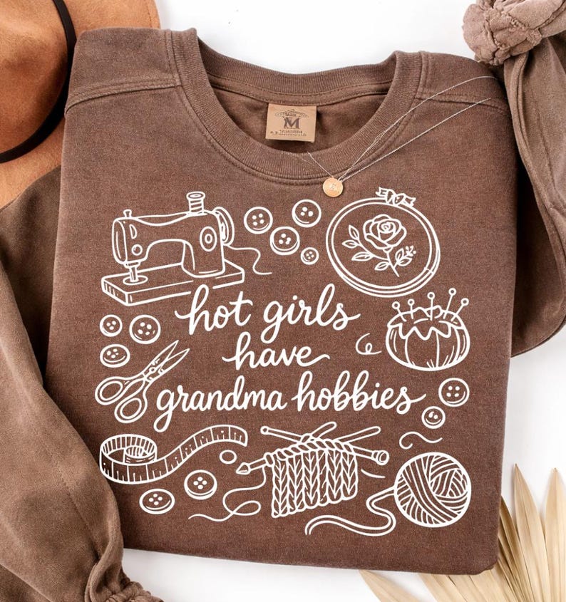 May include: A brown sweatshirt with white graphic illustrations of sewing tools, buttons, and knitting supplies. The text "hot girls have grandma hobbies" is written in a cursive font. A gold necklace and a brown hat are also visible.