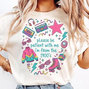 May include: Cream-colored t-shirt with a colorful retro design. The graphic includes a pink star, a boombox, roller skates, and the text "please be patient with me I'm from the 1900's". Other elements include a cassette tape, sunglasses, and a jacket.