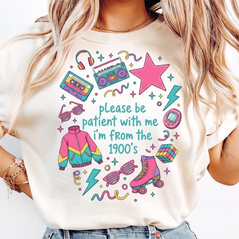 May include: Cream-colored t-shirt with a colorful retro design. The graphic includes a pink star, a boombox, roller skates, and the text "please be patient with me I'm from the 1900's". Other elements include a cassette tape, sunglasses, and a jacket.