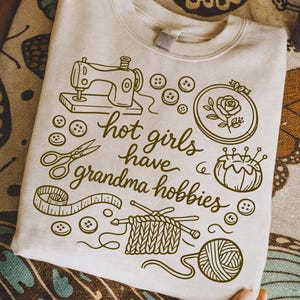 May include: Beige sweatshirt with a brown graphic design featuring a sewing machine, scissors, buttons, and the text "hot girls have grandma hobbies." Other illustrations include a rose in a hoop, a pincushion, knitting needles, and a ball of yarn.