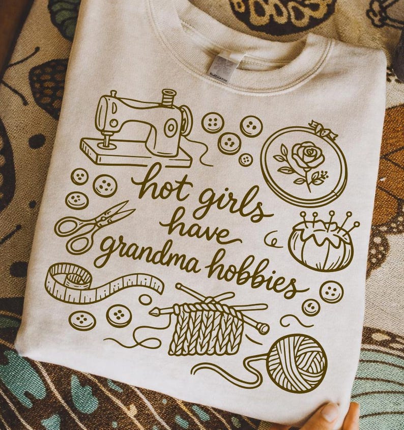 May include: Beige sweatshirt with a brown graphic design featuring a sewing machine, scissors, buttons, and the text "hot girls have grandma hobbies." Other illustrations include a rose in a hoop, a pincushion, knitting needles, and a ball of yarn.